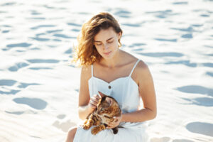 Cute girl in white dress on the beach hugs her bengal cat